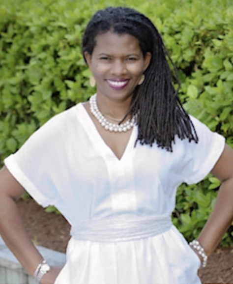 A woman with long, dark, braided hair wears a white dress and pearl jewelry, smiling outdoors in front of green bushes.